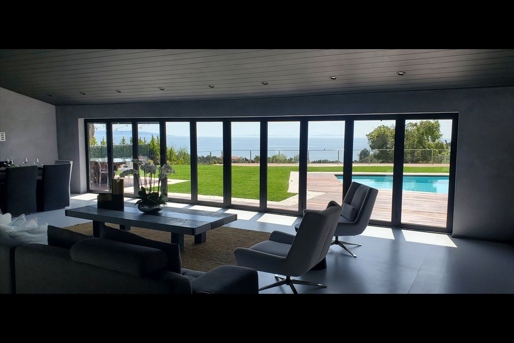 Spacious luxury living room with full-width bifold glass doors opening to a lawn, pool and sea view.